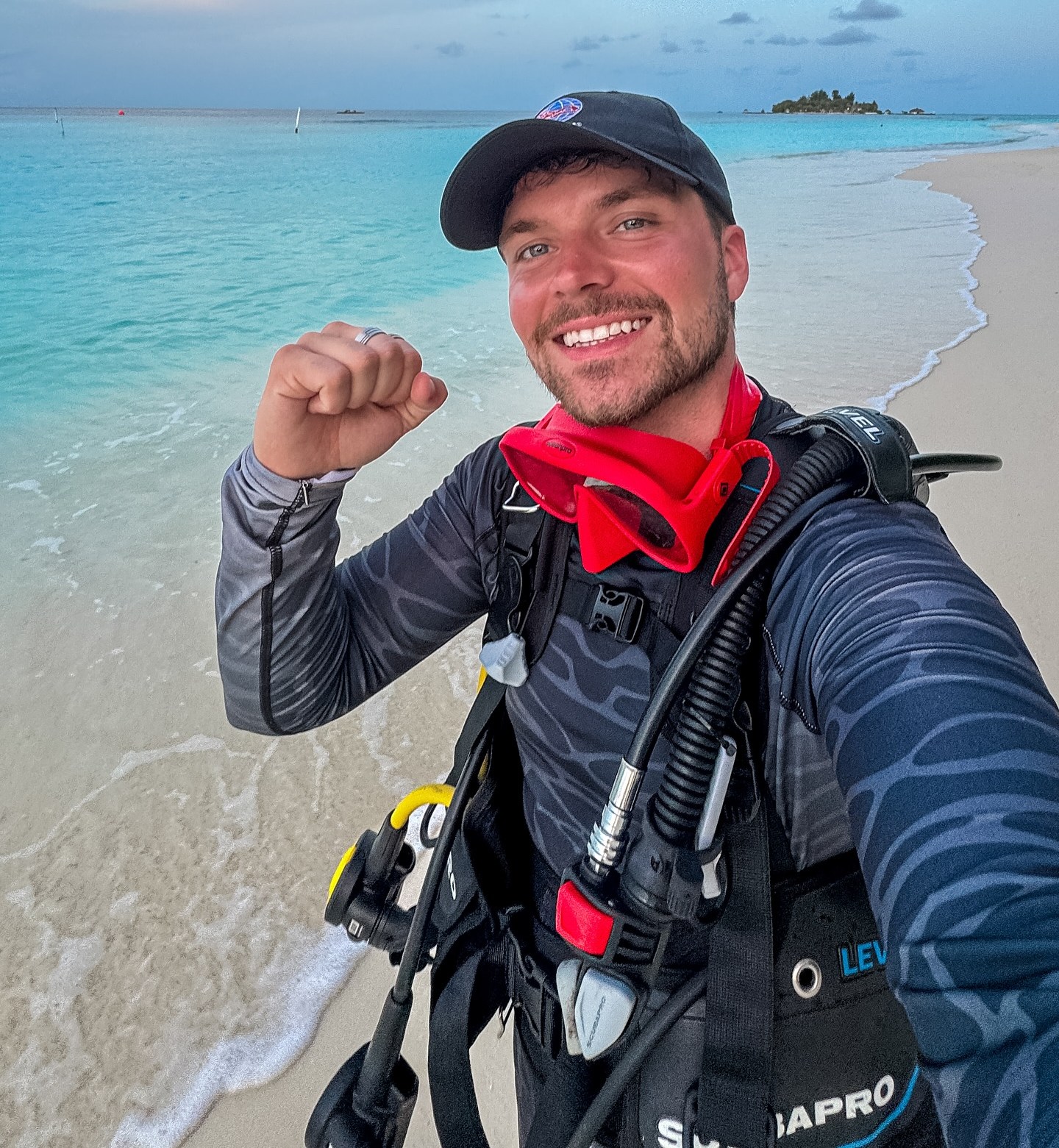 Cam Whitnall, wearing scuba gear, flexing his arm on a sandy beach next to turquoise water.
