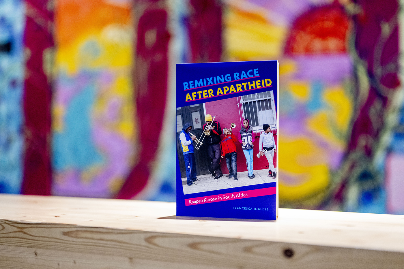 The book "Remixing Race After Apartheid, with a mostly blue cover with a photograph of street musicians, is presented against a very colorful wall.