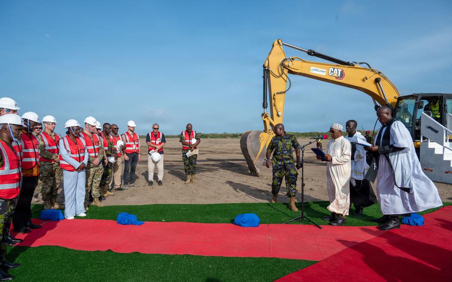 A groundbreaking ceremony with a large yellow CAT excavator positioned next to a red carpet. On the left, a group of officials and service members wearing white hard hats and red safety vests stand watching.