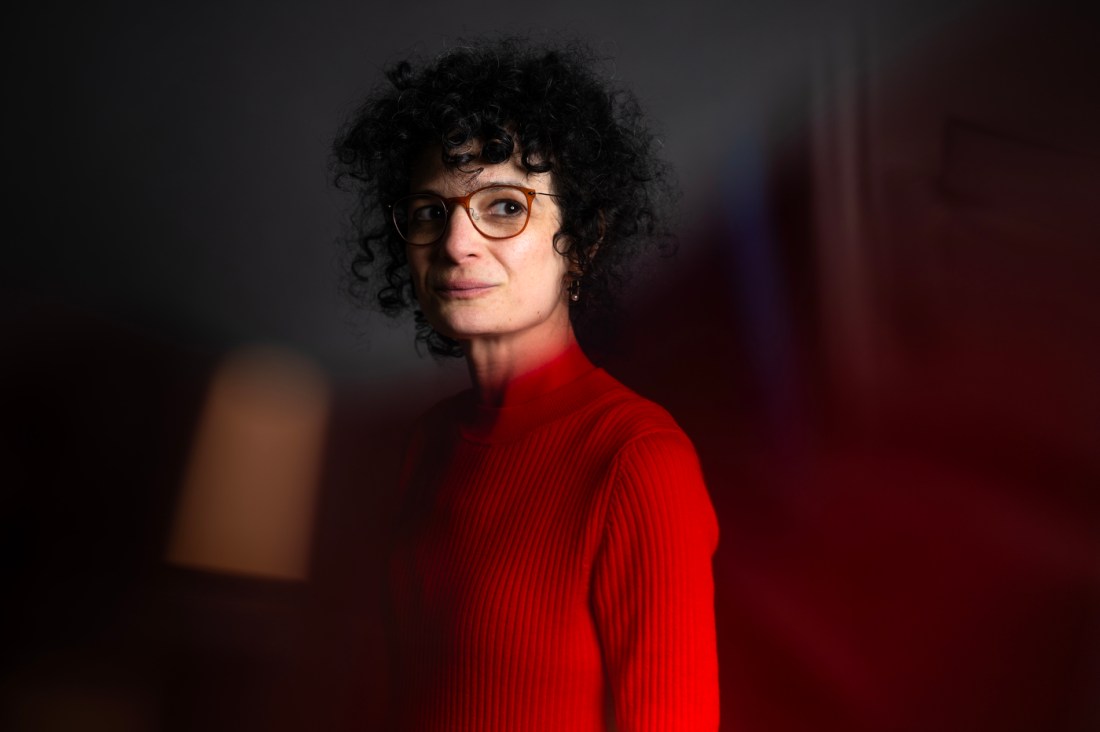 A woman wearing a red turtleneck stands against a dark background. She has orange glasses and curly hair.
