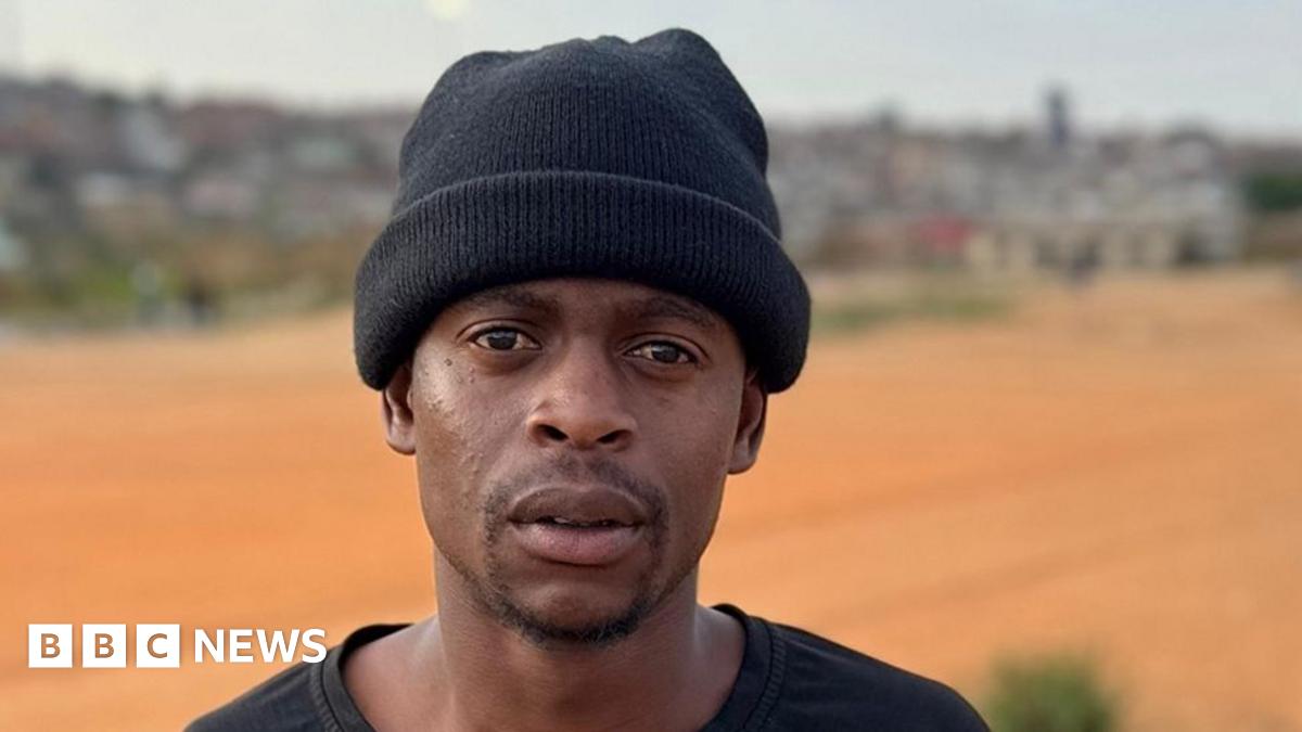 A shot of Arnold Ncube from the waist up. He is wearing a grey V-neck vest and black beanie hat. His right arm is slightly blurred as he can be seen wiping a car.