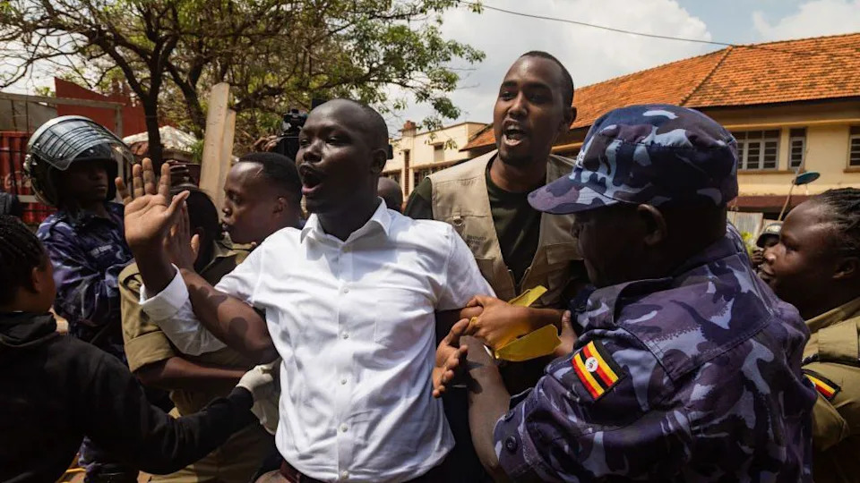 A man in a shirt holds his arm up and exclaims while being restrained by people in uniform