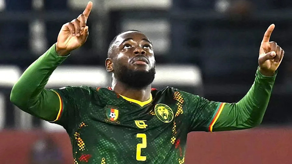 Cameroon full-back Junior Tchamadeu seen from waist up celebrating a goal against South Africa at Afcon 2025 by raising his two arms, with elbows bent, so he can point upwards with his index fingers. He is also raising his head to look up to the sky. Tchamadeu is wearing a green Cameroon jersey with yellow trim and a yellow number two on the centre of the chest, with long green under-sleeves