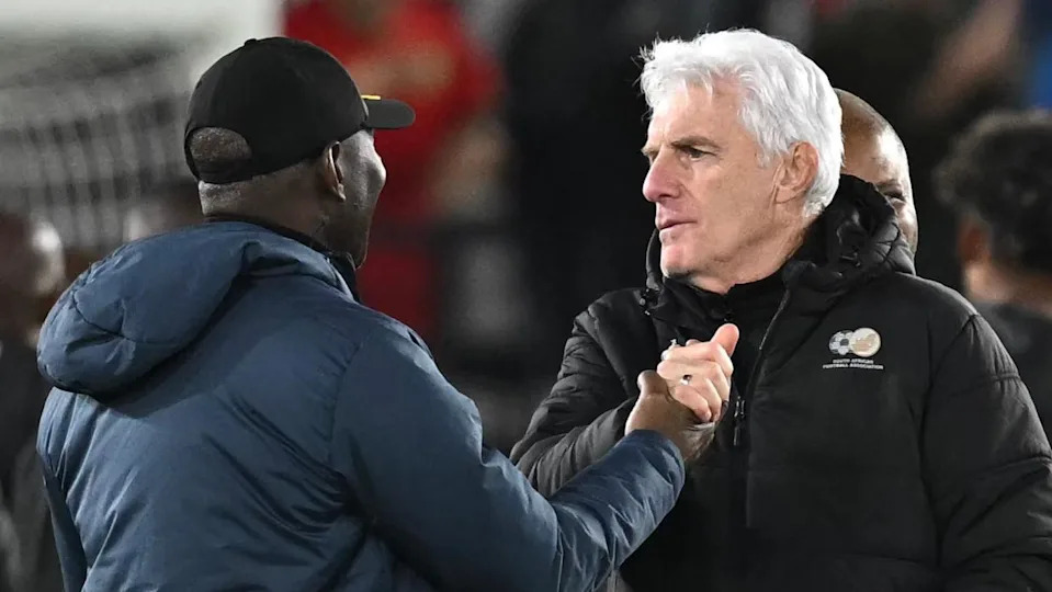 Hugo Broos, wearing a black South Africa puffer jacket, grasps hands with a member of Cameroon's coaching staff, who is seen from the rear