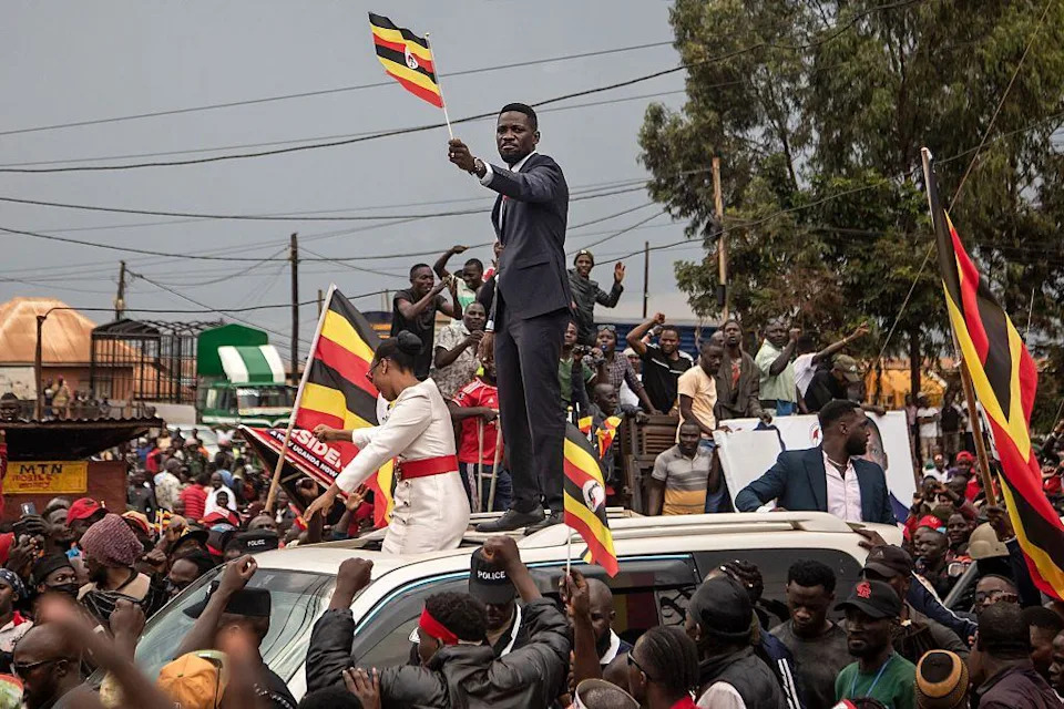 Wine stands atop a car, which is crowded by supporters. Wine and the supporters wave Uganda flags.
