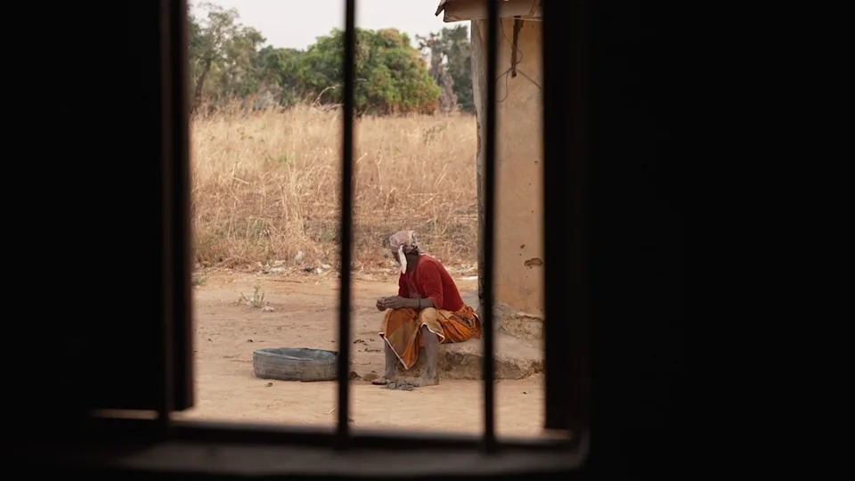 Sarah Peter sitting on a concrete slab. She is seen in the distance and photographed through the bars on a window. She is turning away from the camera.