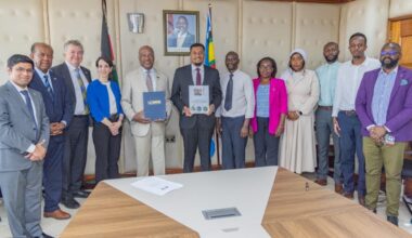 12 people dressed in business attire stand and face camera around a board room table. Two men in the center hold up folders and a document.