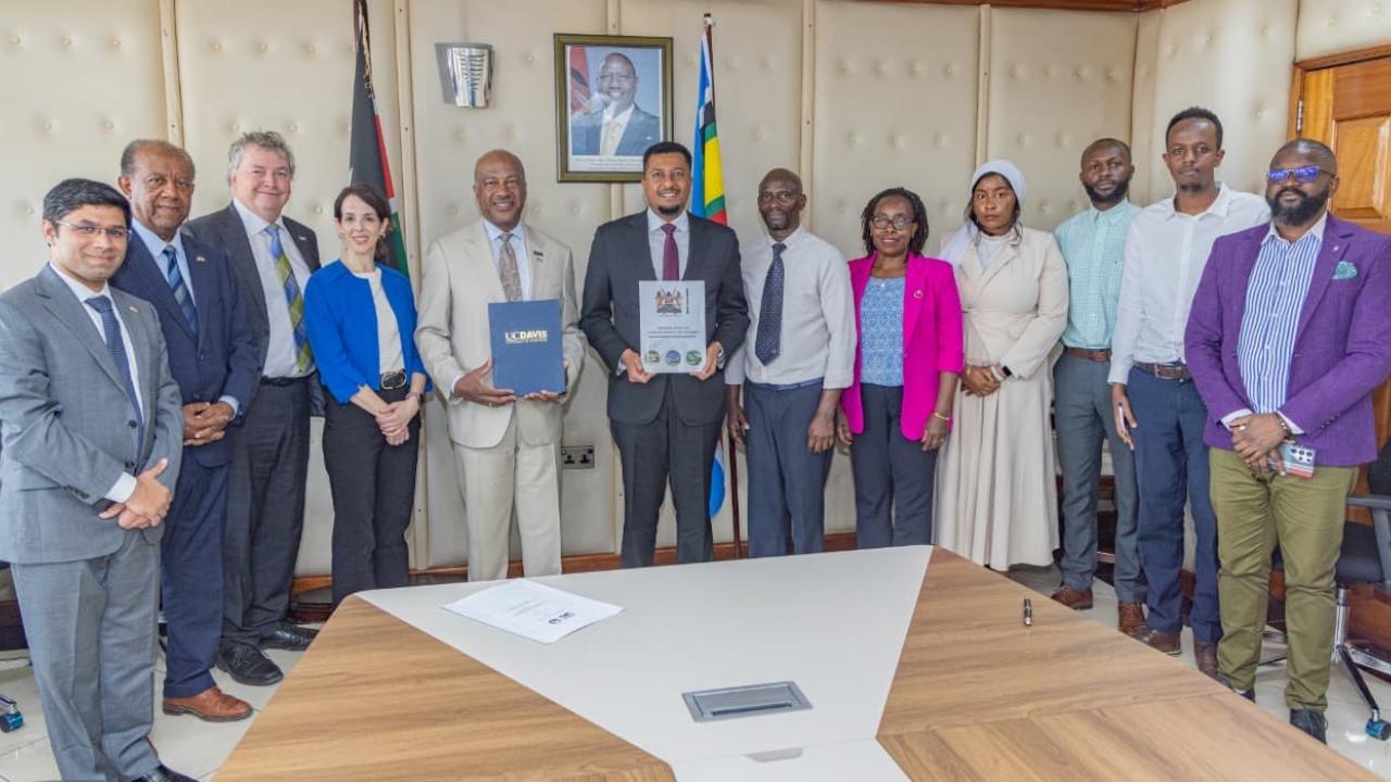 12 people dressed in business attire stand and face camera around a board room table. Two men in the center hold up folders and a document.