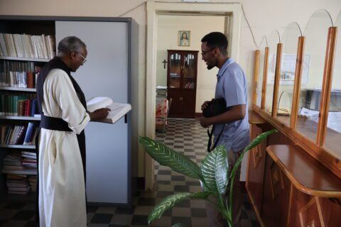 An older man in religious attire reads a book near shelves, while a younger man with a camera listens. The room has a peaceful, studious atmosphere.