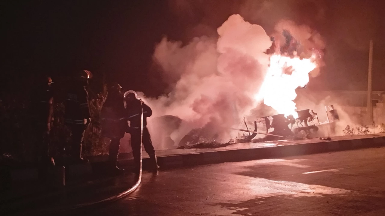 Firefighters with a hose spray a burning truck that is engulfed in flame and smoke and has left the pavement, at night.