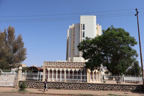 A tall, white modern building stands against a clear blue sky, with a lush green tree and a stone fence in the foreground. A person walks along the sidewalk, lending a sense of scale.