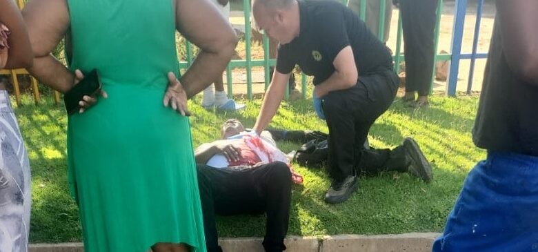 A person lies on the grass, receiving medical attention from a responder, surrounded by concerned onlookers near a colorful fence, conveying urgency and concern.