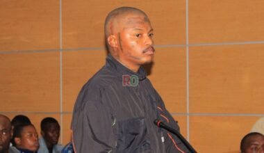 Close-up of Rapper Shebeshxt standing in a courtroom, looking to the side, wearing a black jacket, with visible face and head tattoos.