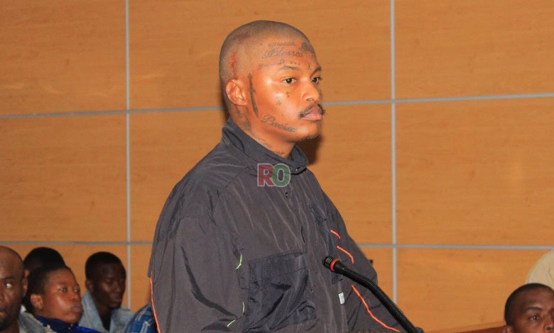 Close-up of Rapper Shebeshxt standing in a courtroom, looking to the side, wearing a black jacket, with visible face and head tattoos.