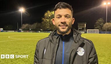 Macauley Bonne at Maldon and Tiptree FC's ground. He wears winter football gear and the pitch is floodlit at night. He has short dark hair and a neat beard and moustache.