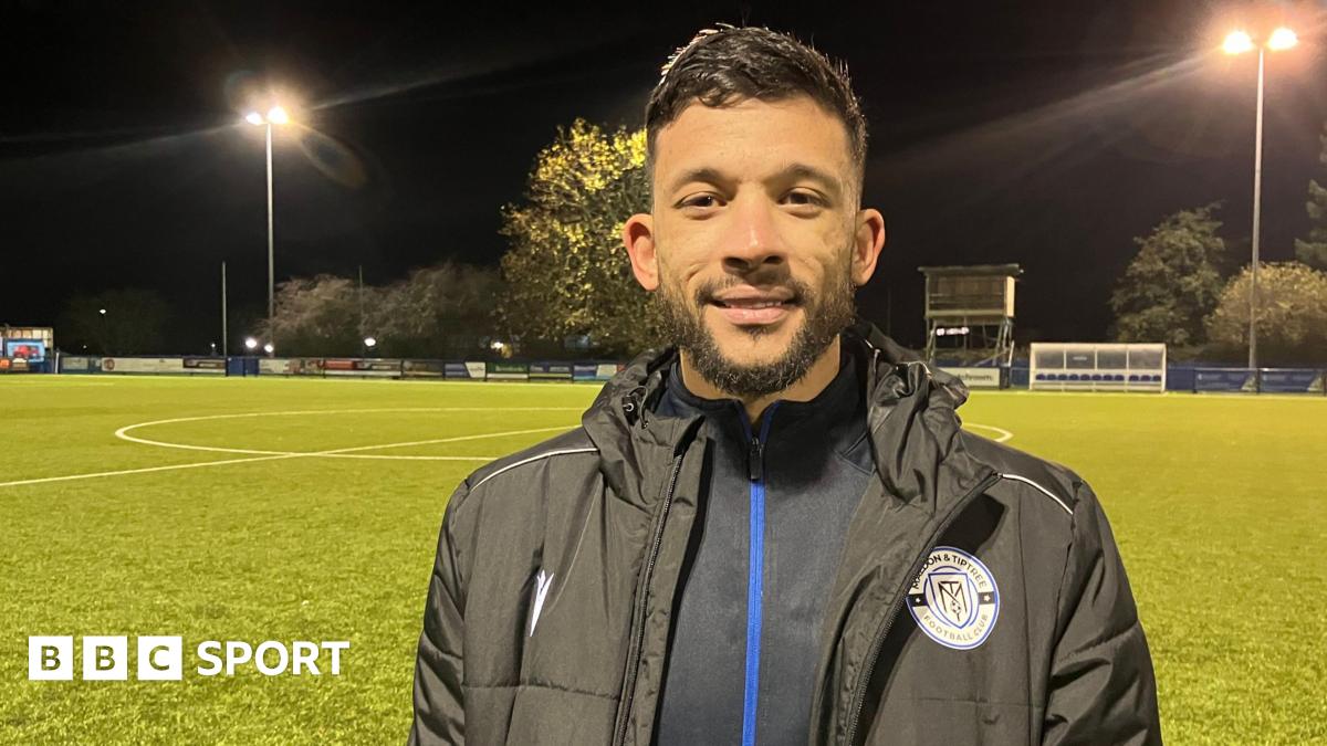 Macauley Bonne at Maldon and Tiptree FC's ground. He wears winter football gear and the pitch is floodlit at night. He has short dark hair and a neat beard and moustache.