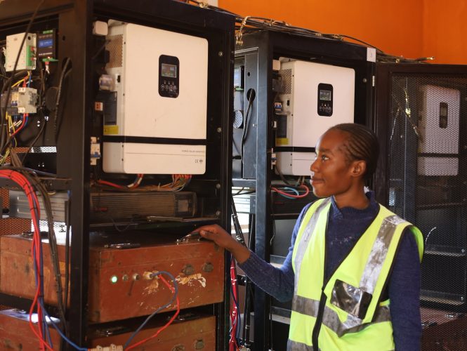 Vimbai Kazembei wears a high visibility vest and stands next to a row of shelves holding e-tricycle batteries connected to chargers. 