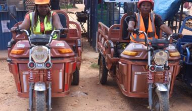Rudo Munguma and Christine Mutsveta sit on large, brown, cargo e-tricycles near stands at the market.