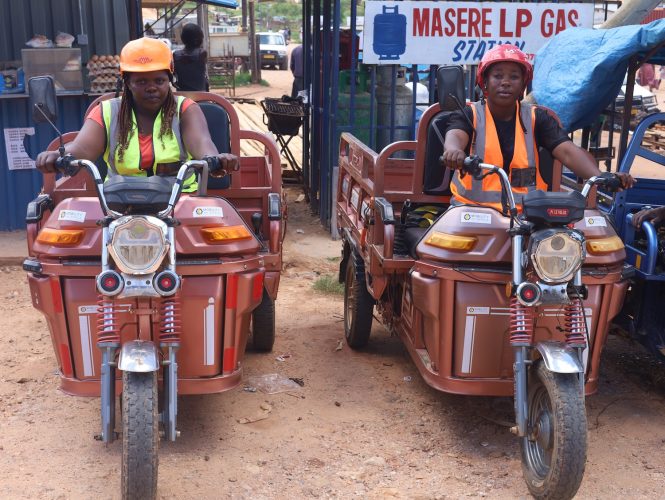 Rudo Munguma and Christine Mutsveta sit on large, brown, cargo e-tricycles near stands at the market.