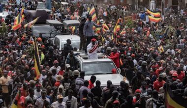 Robert Kyagulanyi, also known as Bobi Wine, addresses supporters from the roof of a car amid a large crowd waving Ugandan flags.