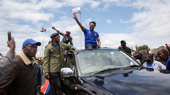 Venâncio Mondlane stands through a car sunroof, raising a hand in a peace sign as supporters gather around during a protest.