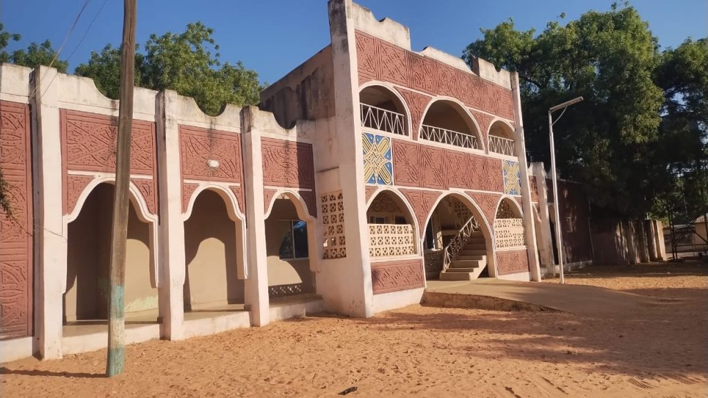 Ornate building with arches and decorative patterns on a sandy ground, shaded by trees.