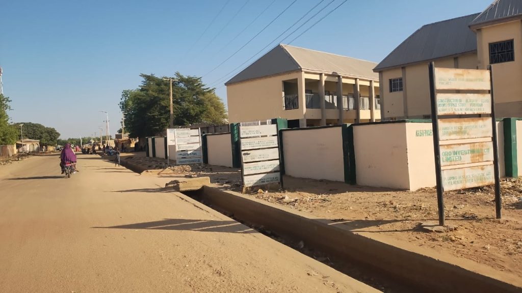 A dirt road with a motorbike and buildings on the right under a clear blue sky, surrounded by signs with text.