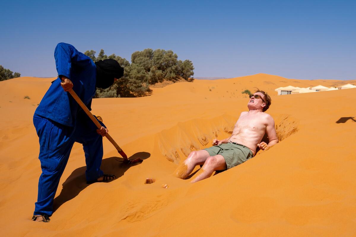 A shirtless man sunbathes on a stretch of sand while another man digs with as shovel nearby.