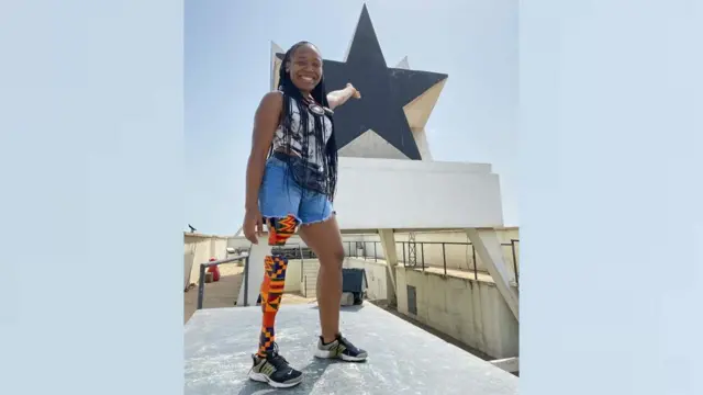 Abena Christine Jon'el debout sur une estrade, avec une prothèse à la jambe droite enveloppée dans un tissu kente. Elle pointe du doigt derrière elle, vers le sommet du monument Black Star d'Accra.