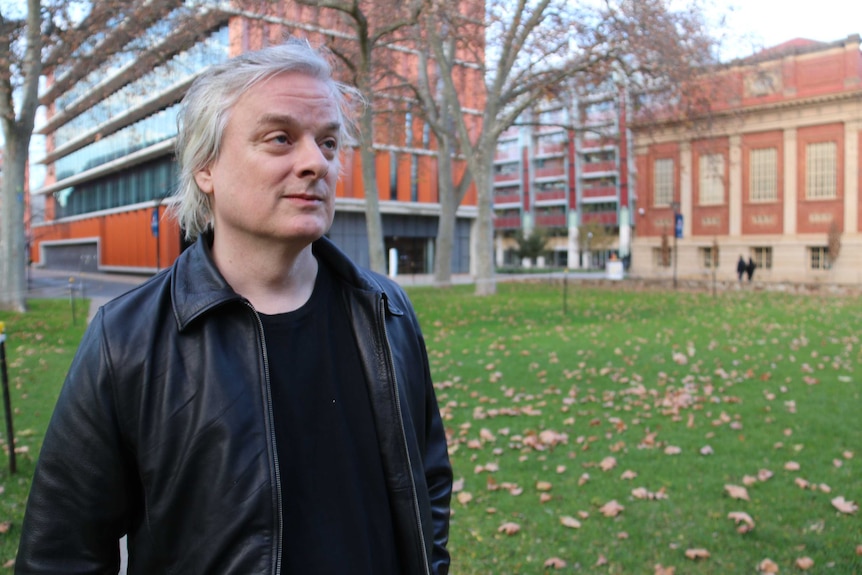 David with black jacket and shirt stands in front of university buildings and a lawn.
