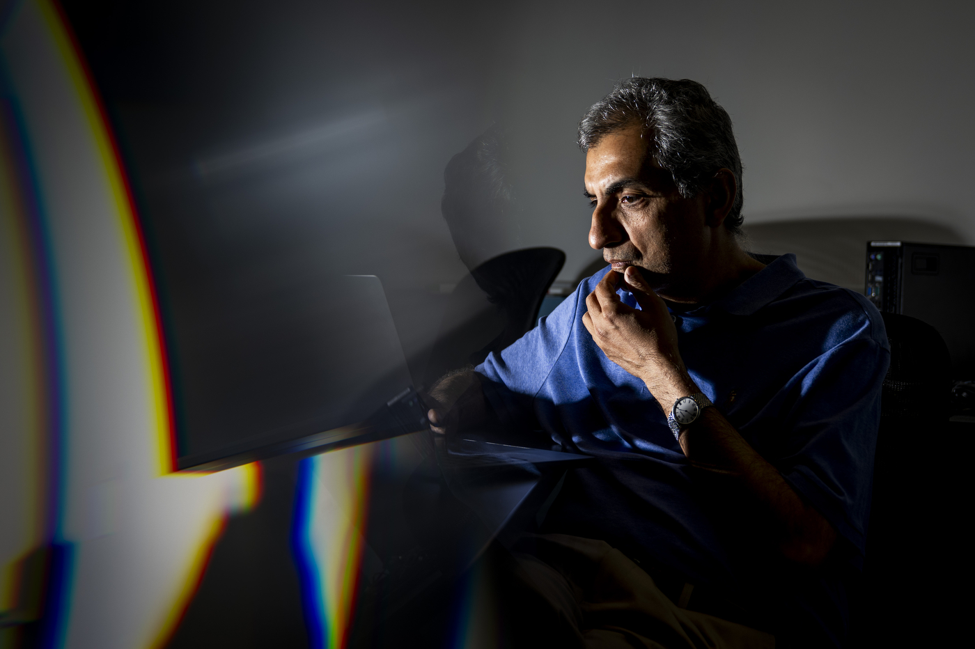 Hossein Mosallaei looks at a laptop screen, his face lit from the side against a dark room, with a prism-like streak of rainbow light crossing the foreground.
