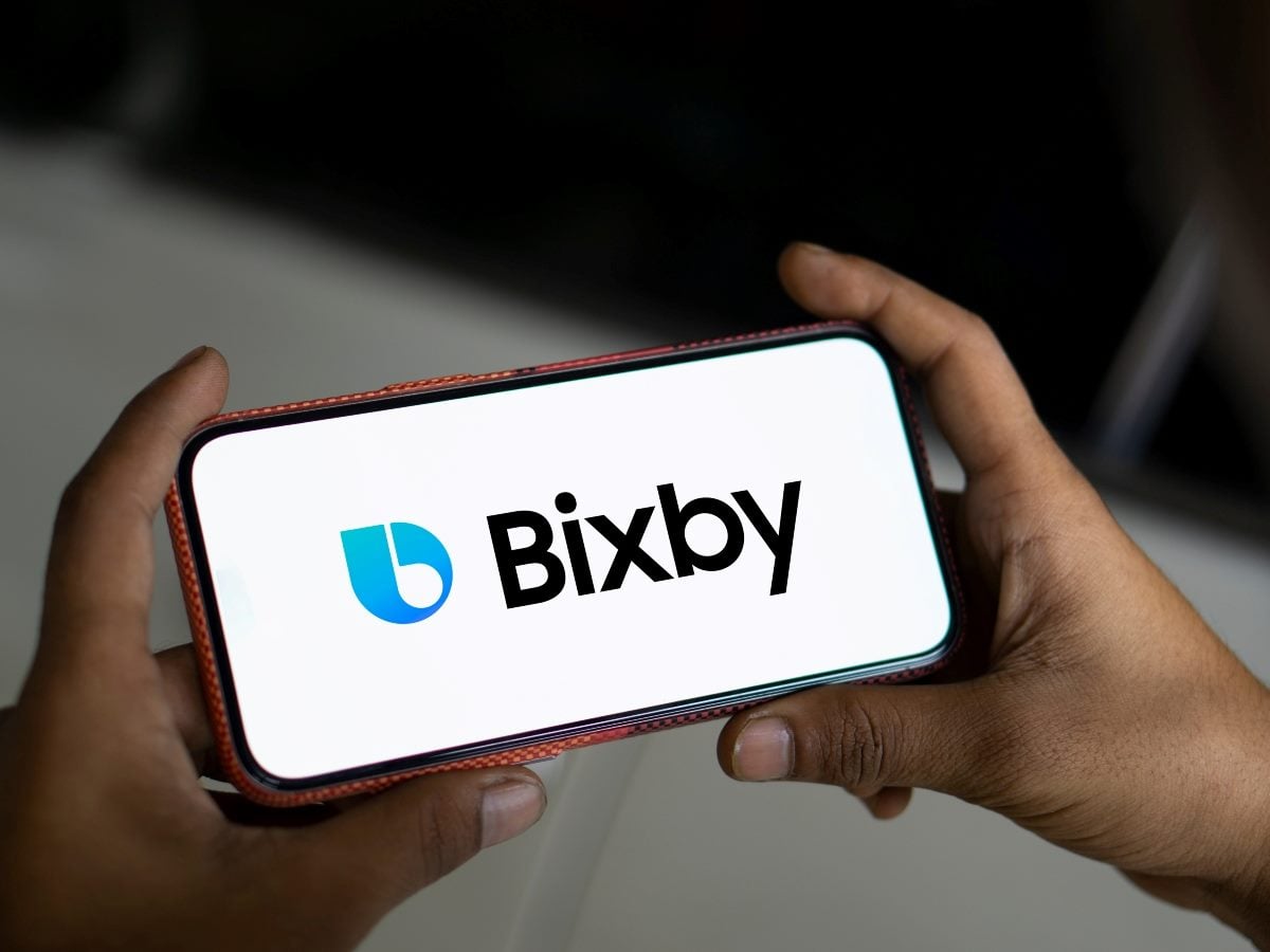 A person's hands holds a smartphone in a red patterned case with both hands against a dark background, displaying the Bixby logo and wordmark in blue and black on a white screen, illustrating Samsung's voice assistant referenced in the article's South Korea example.