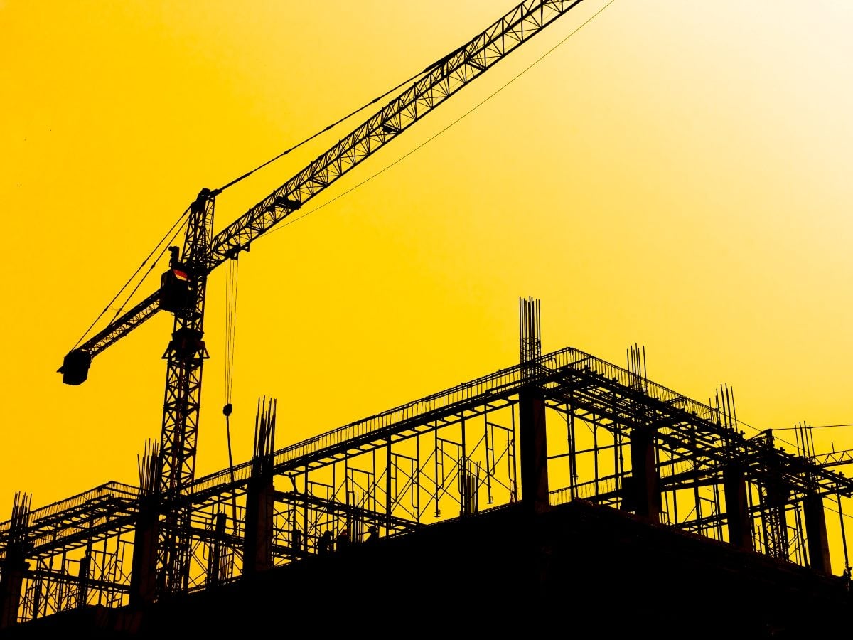 A silhouetted construction crane and multi-story building framework with exposed rebar and scaffolding set against a bright yellow-orange sunset sky, evoking the idea of infrastructure being built from the ground up.