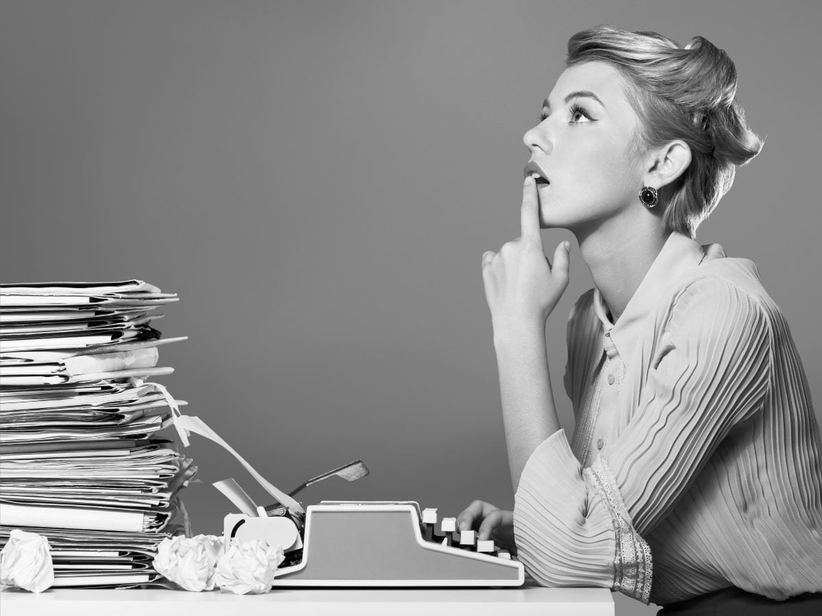 Black-and-white image of a woman seated at a typewriter, pausing thoughtfully beside a large stack of papers and crumpled drafts.