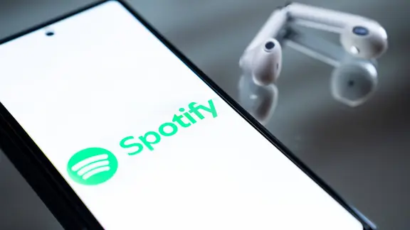 Ein Handybildschirm auf dem das Logo Spotify zu sehen ist, daneben liegen Kopfhörer.