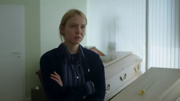 Schauspielerin Magdalena Laubisch im Film "Sechswochenamt". | Filmweh, Markus Ott Eine Frau mit blonden Haaren steht mit verschränkten Armen und entsetztem Blick vor Holzsärgen.