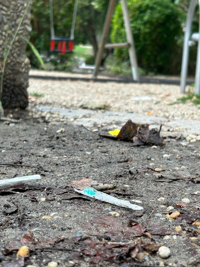 das Bild zeigt Spritzen, die am Erdboden liegen. Im Hintergrund erkennt man den Kinderspielplatz.