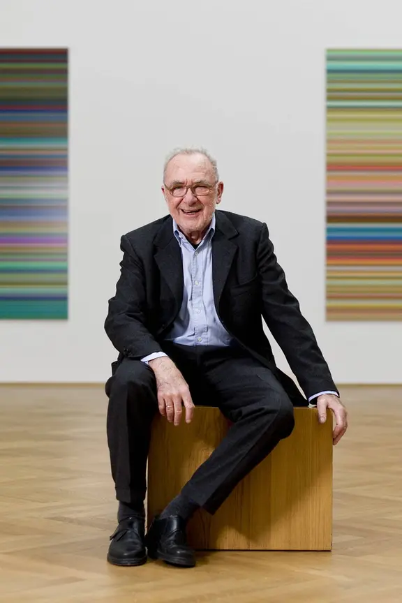 Gerhard Richter in einer Ausstellung im Dresdner Albertinum im Jahr 2013. | IMAGO/Robert Michael Ein als der Maler Gerhard Richter bekannter älterer Mann sitzt in einem dunklen Anzug in einer Kunstgalerie auf einem gelben Würfel.