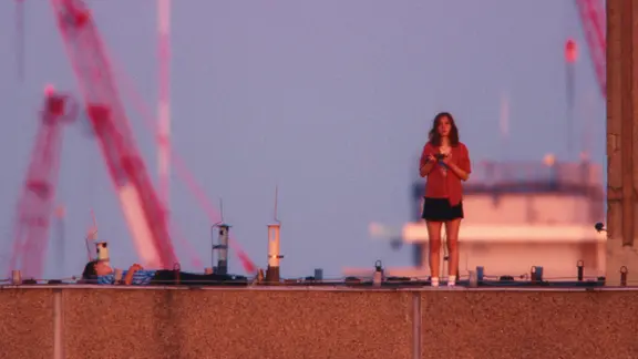 Szene aus dem Film "Everytime": Eine junge Frau steht auf einem Flachdach, während ein junger Mann links auf dem Dach liegt. | TheBarricades, PanamaFilm, Gregory Oke Szene aus dem Film "Everytime": Eine junge Frau steht auf einem Flachdach, während ein junger Mann links auf dem Dach liegt.