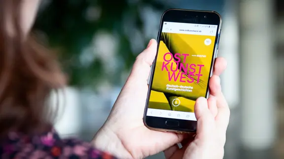 Eine Frau hält ein Smartphone in der Hand, auf dem Display steht >>ostKUNSTwest<< | MDR/Robert Hensel Eine Frau hält ein Smartphone in der Hand, auf dem Display steht >>ostKUNSTwest<<