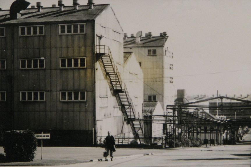A black and white photo of a person walking away from a large multi-storey building with stairs leading almost to the top.