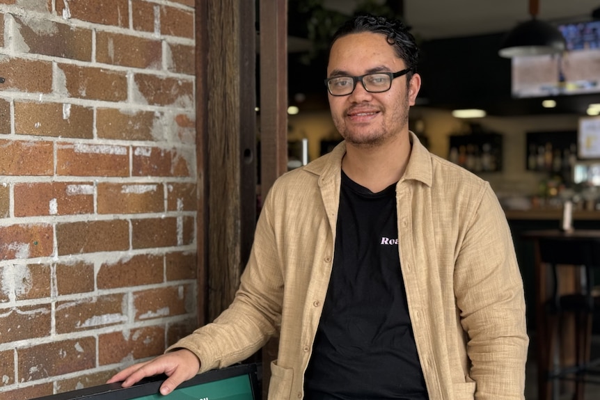 A man in a tan button shirt and black tee smiles at the camera