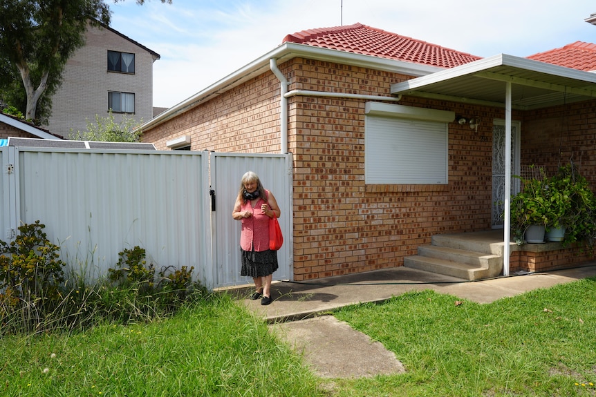 Lily stands out of the front of her brick home and in front of grass, white doors, red roof.