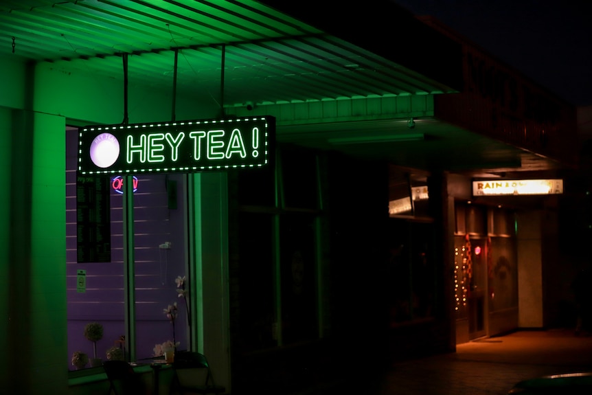 A street scene in the dark a shop in the foreground is lit up in green and has a neon sign reading HEY TEA, hanging in front.