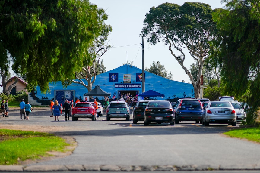 People walk past parked cars outside a blue brick building that says "Rosebud Sea Scouts".