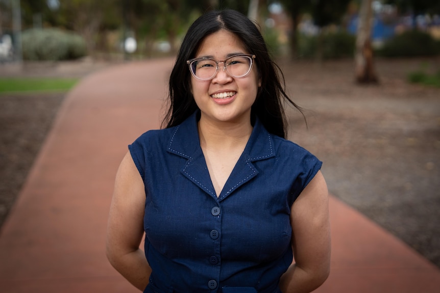 A woman with black hair and glasses stands with her hands behind her back smiling.