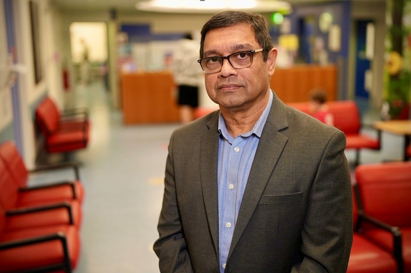 A close up of a man with glasses wearing a suit and standing in a hospital room.