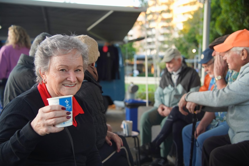 A woman smiles and holds up a coffee while sitting with a group of people.