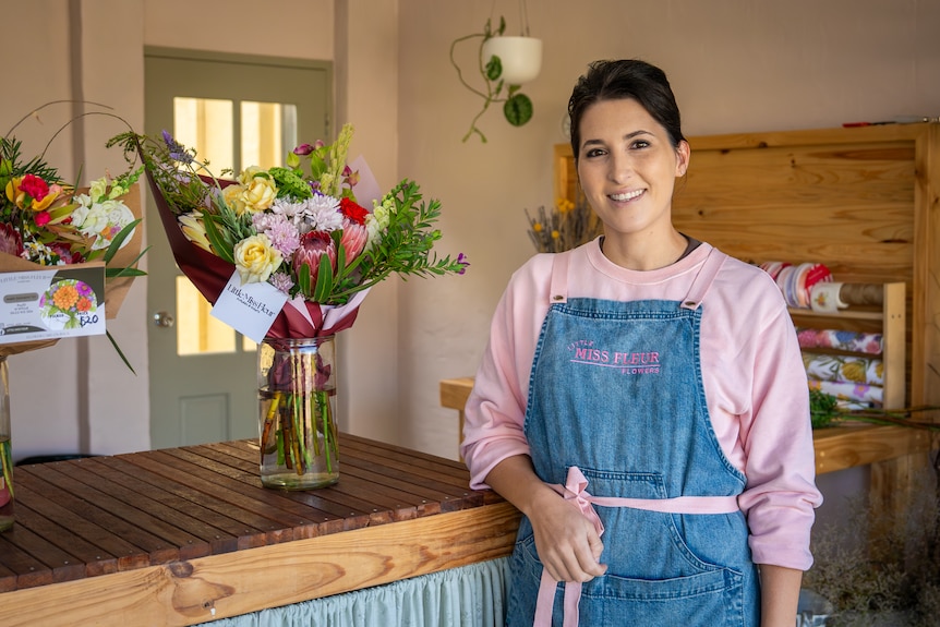 A woman has dark her and a pink jumper, she stands leaning on a wooden bench with a bunch of colourful flowers