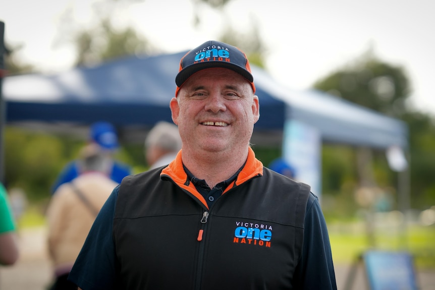A man wearing a jacket and hat reading One Nation smiles for the camera.